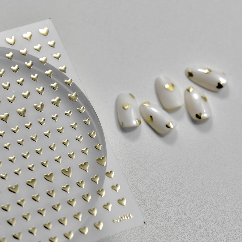 Gold heart 3D nail art stickers shown with white sample nails featuring metallic heart accents.